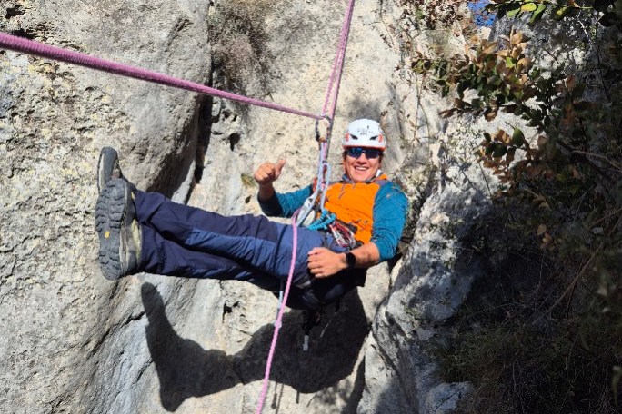 Person rock climbing with ropes, wearing a helmet and orange vest, in a rocky outdoor setting.