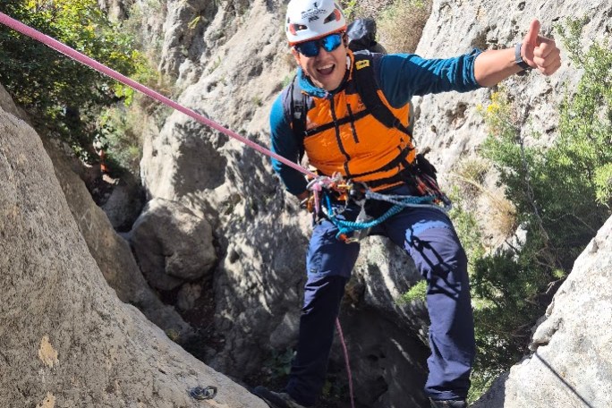 Climber in gear giving thumbs up while rappelling on a rocky cliff.