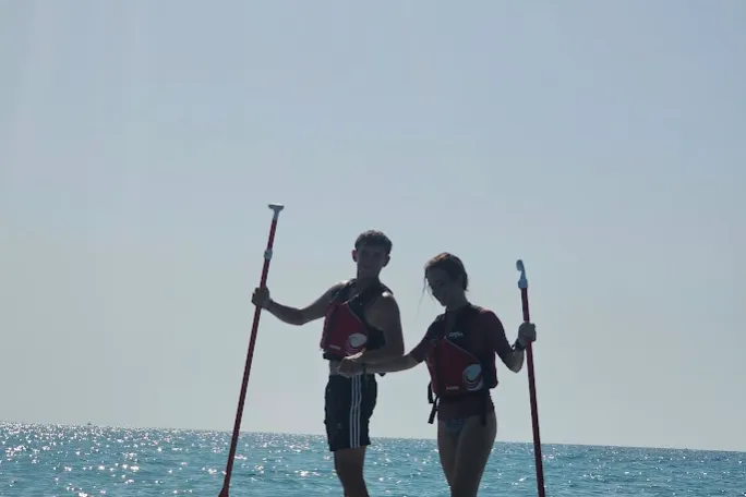 Two people stand on a paddleboard in the sea holding paddles, silhouetted against the sky.