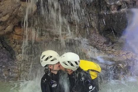 Two people in helmets embrace under a waterfall in an outdoor setting.