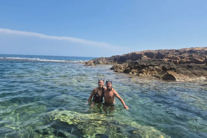 Two people standing in clear ocean water near a rocky shoreline under a blue sky.