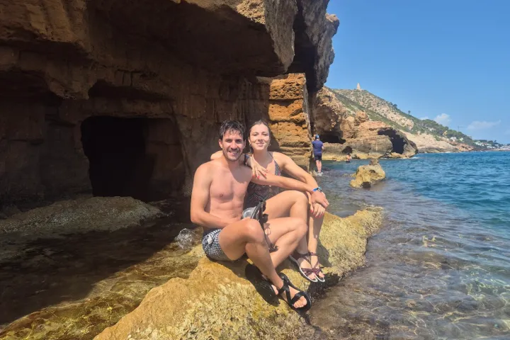 Couple sitting on rocks by the sea with cliffs in the background on a sunny day.
