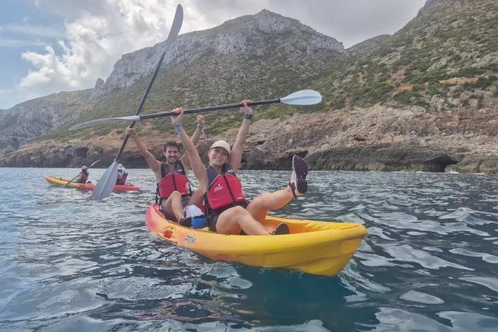 Two people kayaking in a sea near rocky cliffs, smiling and holding paddles overhead.