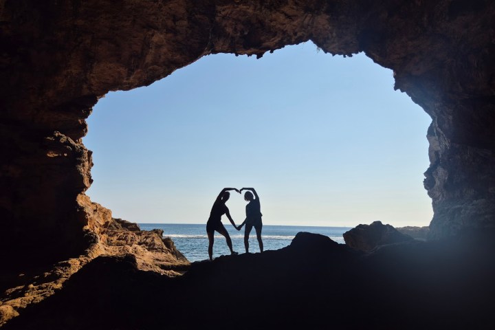 Two silhouetted people forming heart shape in cave opening with sea view.