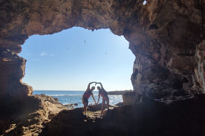 Two people silhouetted forming heart shape in rocky cave with ocean view.