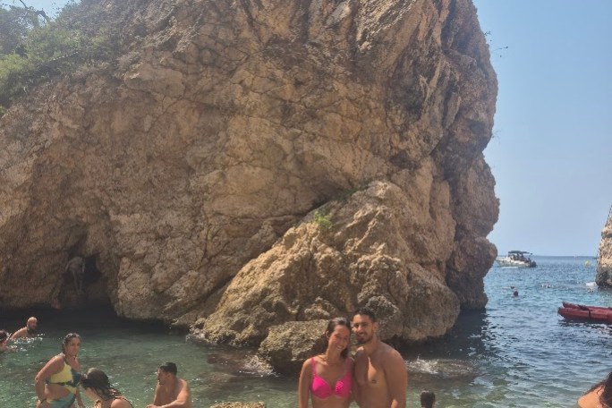 People swimming and standing by rocks in a clear blue sea with a large rocky cliff.