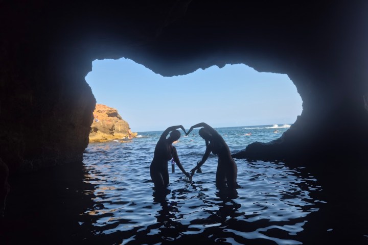 Two people forming a heart shape with their arms in a cave overlooking the sea.