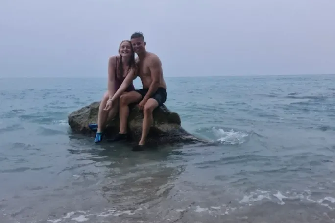 Two people sitting on a rock in the sea, overcast sky in the background.