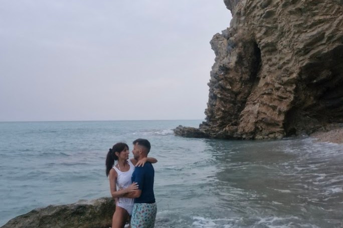 Couple embracing in shallow ocean near a rocky cliff.