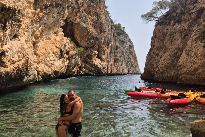 A couple hugs in clear water between rocky cliffs with kayaks nearby.