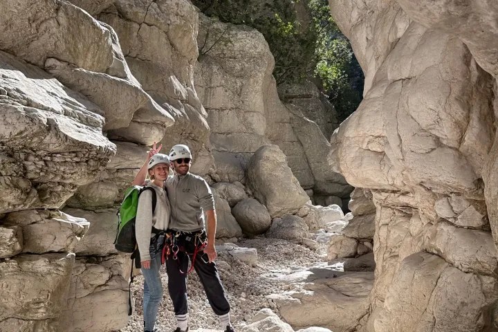 Two climbers in helmets stand in a rocky canyon, surrounded by large boulders.