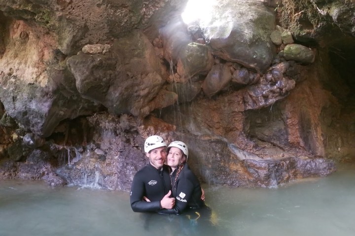 Two people in wetsuits and helmets standing in a rocky cave with water.