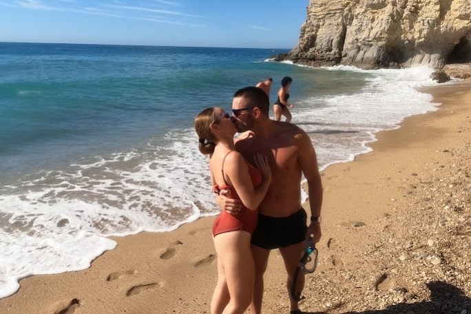 Couple kissing on a sandy beach with a cliff and ocean in the background.