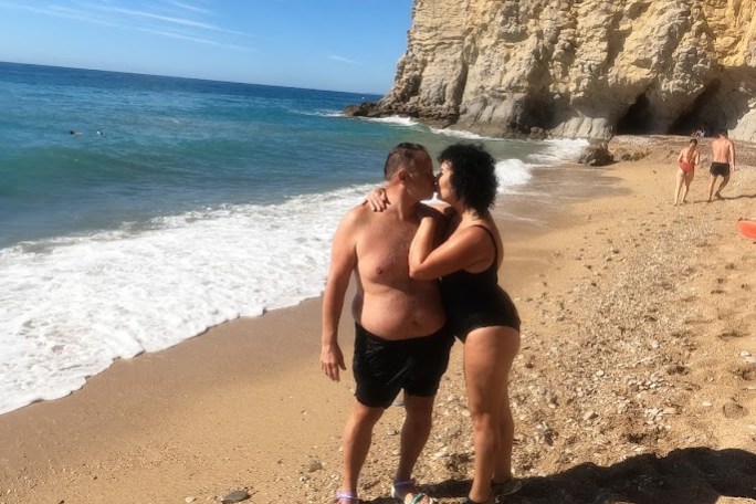 Couple embracing on a sandy beach with a cliff and blue ocean in the background.