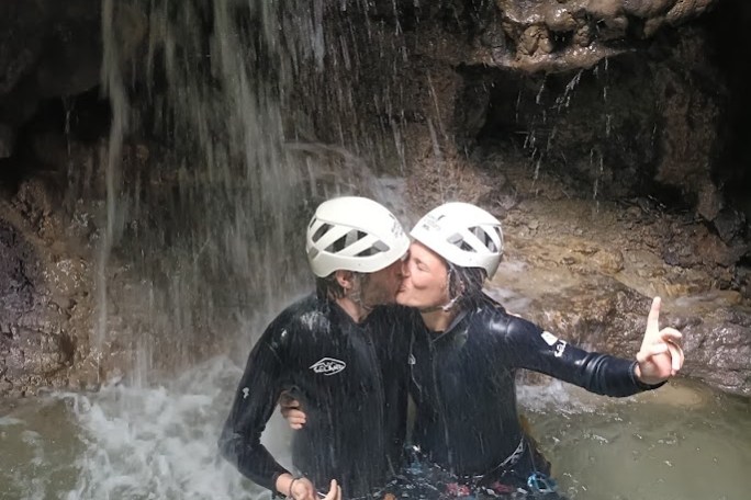 Two people in helmets kissing under a waterfall, standing in water, wearing wetsuits.