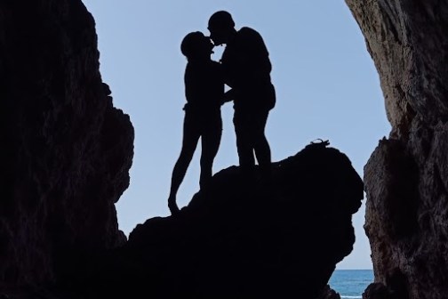 Silhouetted couple kissing in a cave opening with sky background.