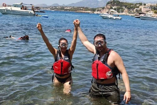 Two people in life jackets and snorkels holding hands in clear water with boats and mountains in the background.