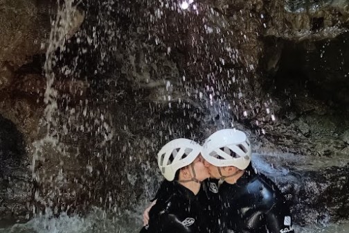 Two people in helmets kiss under a small waterfall in a rocky area.