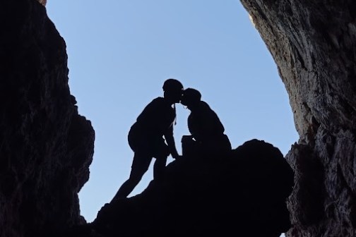 Silhouette of two people kissing in a cave opening with sky visible.