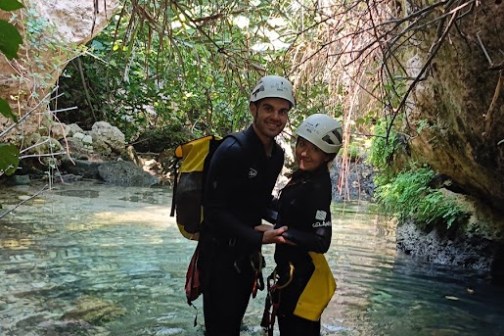 Two people in wetsuits and helmets standing in a shallow stream under hanging vines.