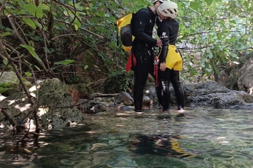 Two people in wetsuits by a forest stream, surrounded by lush green foliage.
