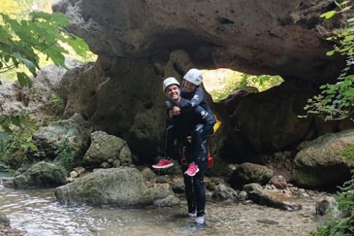 Two people in helmets stand in a shallow, rocky stream under a large, natural rock arch.