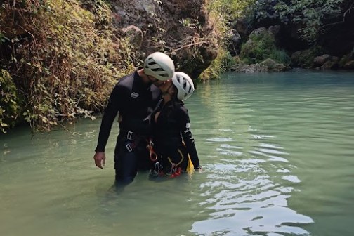 Two people in helmets and wetsuits wading through a forest river.