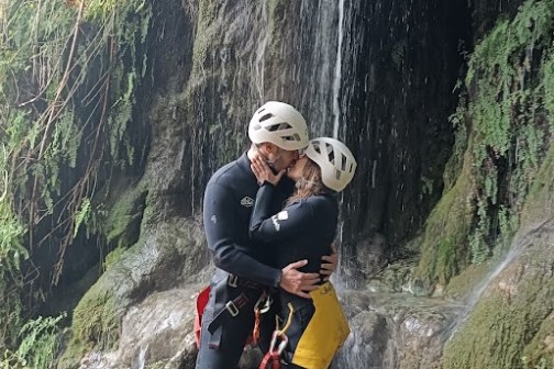 Two people in helmets embrace under a small waterfall.