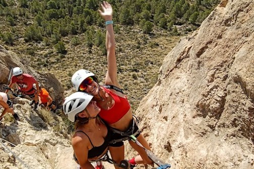 Two climbers on a rocky cliff with helmets and gear, one raising an arm, with a scenic landscape in the background.