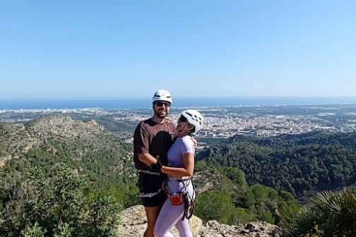 Two people in climbing gear hugging on a rocky hilltop with a view of a city and ocean in the background.
