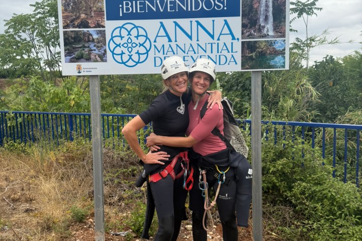 Two people wearing helmets and harnesses, standing and hugging in front of a welcome sign.