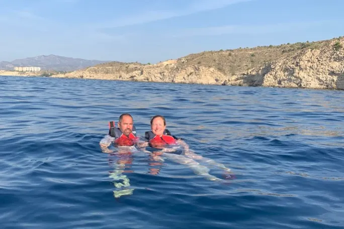 Two people in life vests floating in the ocean near rocky cliffs.