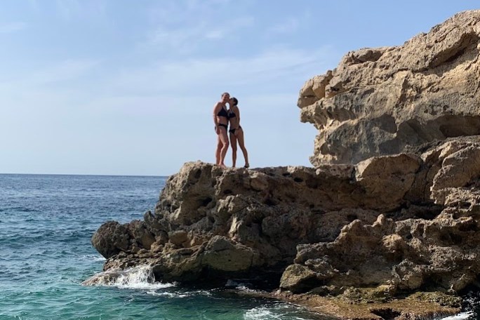 Two people standing on a rocky cliff by the sea, under a clear blue sky.