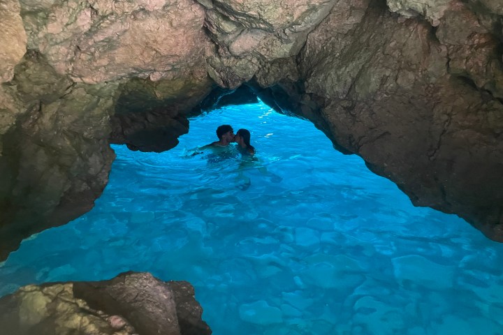 Two people swimming in a blue-lit cave with rocky walls.