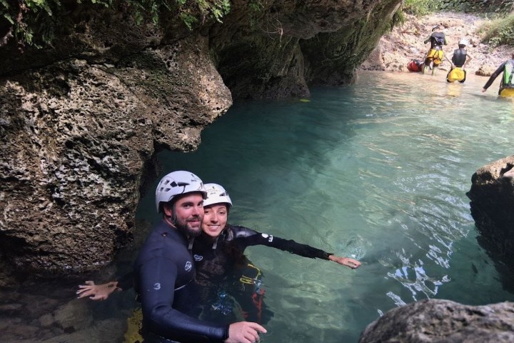 Two people in wetsuits and helmets wading in a rocky clear water canyon with others in the background.