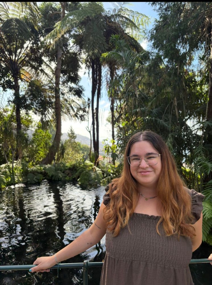 Person with long hair and glasses smiling in front of a lush pond and trees.
