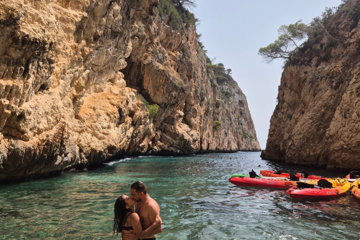 Couple kissing in clear water between tall cliffs, with kayaks floating nearby.