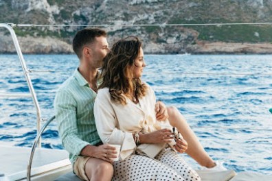 Couple sitting on a boat, gazing at the sea, with rocky coastline in background.