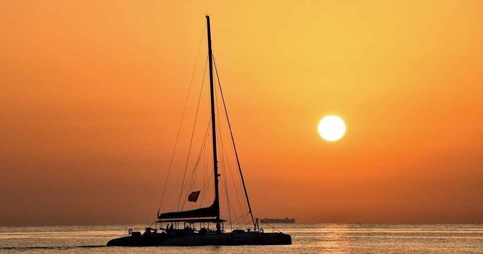 Silhouette of a sailboat on calm sea with an orange sunset in the background.