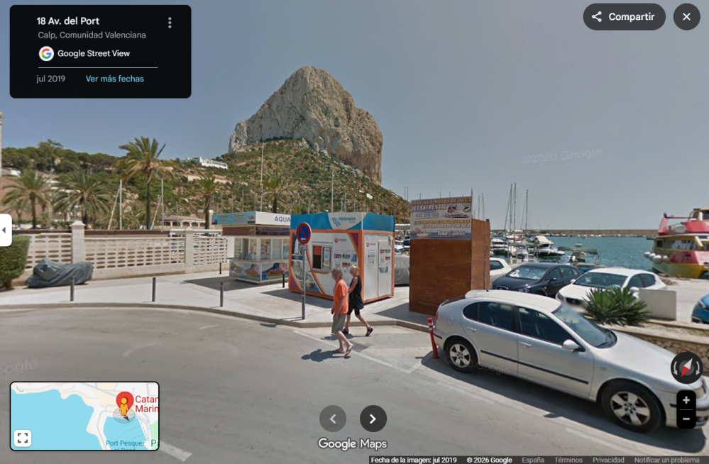 Street view of a coastal area with a large rock formation, parked cars, palm trees, and people walking.