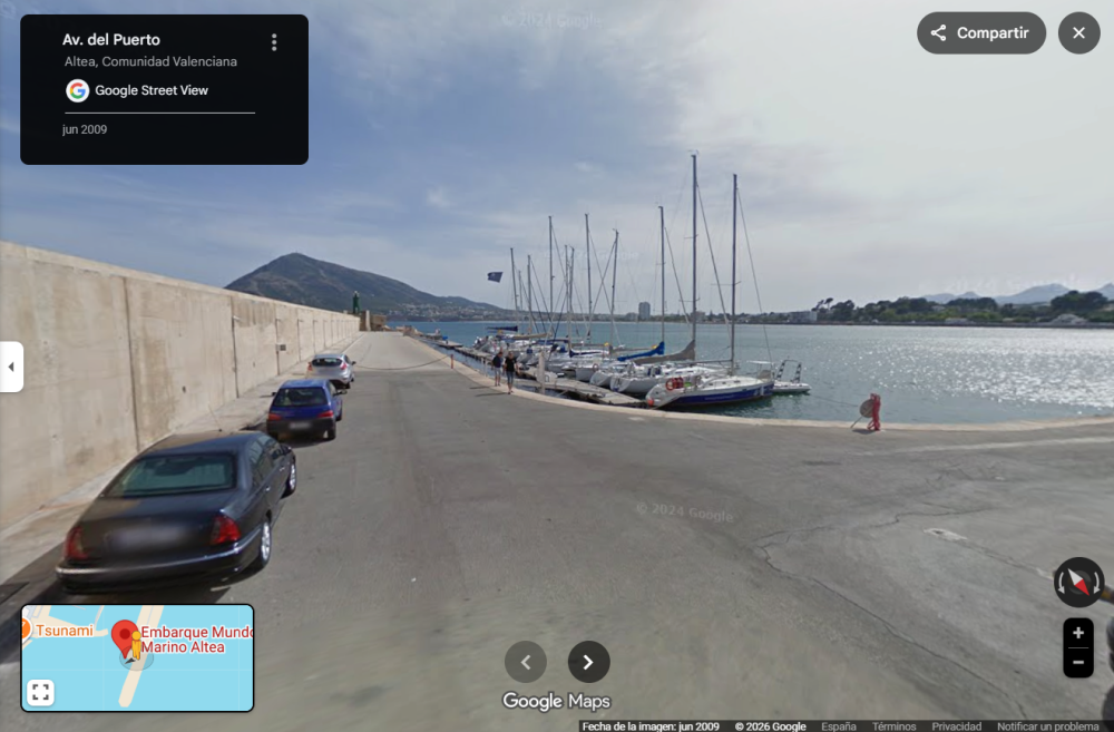 Coastal road with parked cars, sailboats docked at marina, mountains in background.