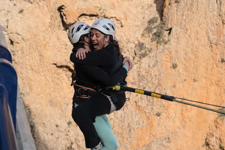Two people in helmets hugging mid-air during a bungee jump near a rocky cliff.