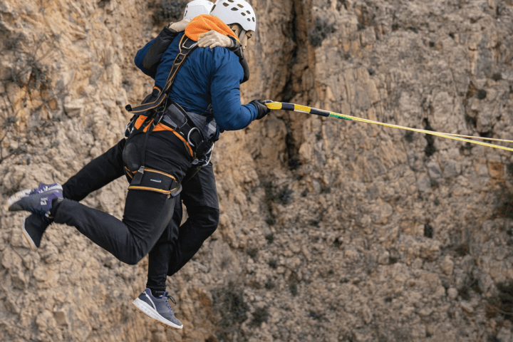 Two people in helmets and harnesses mid-air, rappelling against a rocky cliff background.