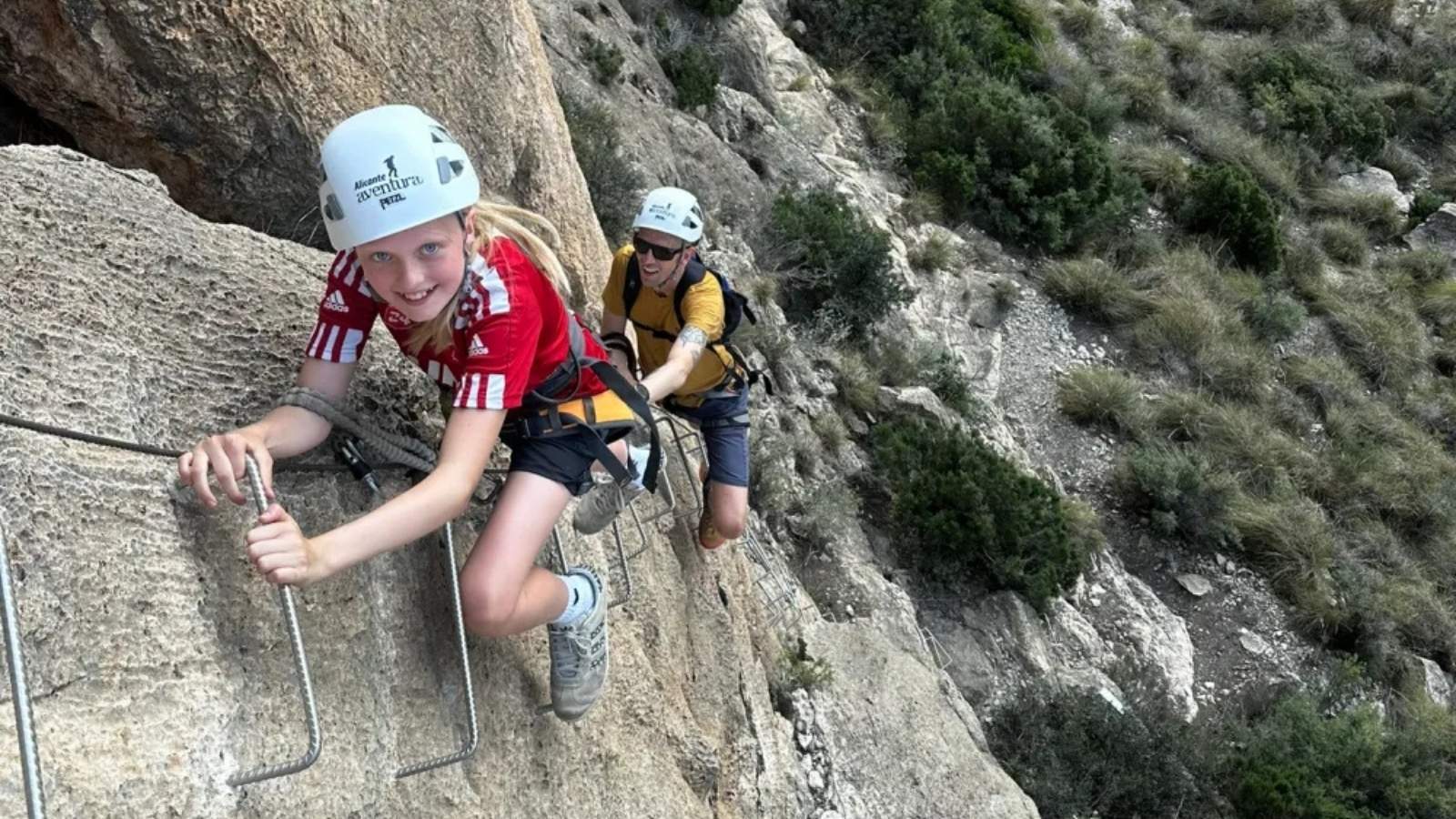 Niña realizando una vía ferrata acompañada por un adulto y equipada con casco y arnés
