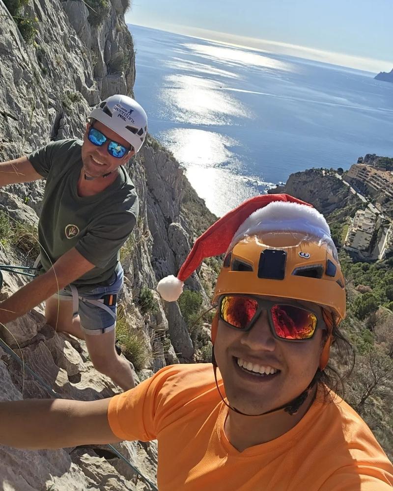 Dos participantes sonrientes con casco y arnés durante un bautismo de escalada en el Morro de Toix, con el mar Mediterráneo de fondo